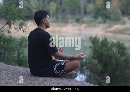 Un jeune homme est assis paisiblement au bord d'un lac tranquille, méditant dans un état calme et réfléchi. La scène sereine capture l’harmonie entre le corps et la nature Banque D'Images