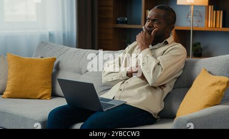 Réfléchi perplexe homme d'affaires afro-américain freelance travaillant sur ordinateur portable à la maison canapé penser à résoudre le problème d'affaires sérieux pensif Banque D'Images