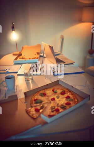 Pizza, ordinateur portable et documents sur table dans le bureau la nuit pour la date limite, la gestion de projet et manger. Restauration rapide, technologie et paperasse sur le bureau à Banque D'Images