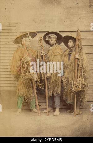 Trois Japonais vêtus de robes d'herbe et de chapeaux à larges bords, 1868-1880. Portrait de trois hommes debout devant un bâtiment. Chacun porte un mino, ou cape de paille pour repousser la pluie, et un chapeau de paille à larges bords. Chaque homme porte également un long poteau avec un filet de corde noué attaché. Banque D'Images