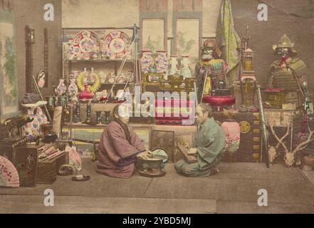 Deux hommes d'affaires japonais dans leur magasin, 1868-1880. Portrait de deux hommes assis dans un magasin face à face. L'un des hommes tient ce qui semble être un boulier, et l'autre repose ses mains sur un bol. Ils sont entourés par les articles à vendre dans la boutique, qui comprend des armures, des bols et des vases en porcelaine, des ventilateurs, des épées et d'autres objets décoratifs. Banque D'Images