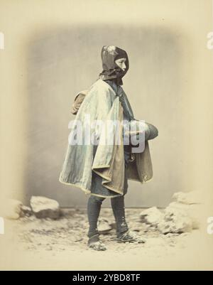 Sujet en robe d'hiver, vers 1868. Une personne se tient contre un fond enneigé portant une capuche et une cape. Une corde avec un paquet enveloppé à chaque extrémité pend sur leur épaule. Banque D'Images