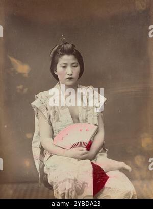 Costume d'été musum, années 1870-1890s. Portrait d'une fille assise portant une robe sans manches. Elle tient un ventilateur et a un pied reposant sur son autre jambe. Banque D'Images