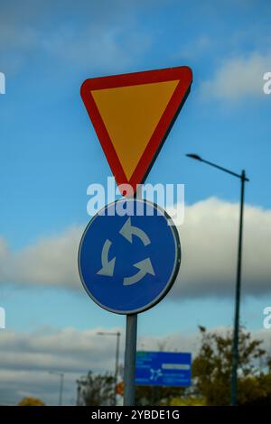 Un panneau de limite jaune triangulaire est placé au-dessus d'un panneau circulaire bleu indiquant un rond-point, placé contre un ciel bleu clair en milieu urbain Banque D'Images