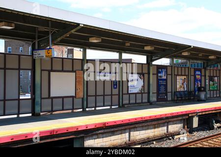 Queens, New York, États-Unis - 20 août 2024 : un après-midi tranquille à la gare de Woodside avec des bancs vides et des panneaux de la gare sous un ciel bleu. Banque D'Images