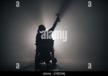 Femme handicapée recevant une coupe de victoire alors qu'elle était assise dans un fauteuil roulant dans une pièce sombre avec un rétroéclairage blanc. Banque D'Images