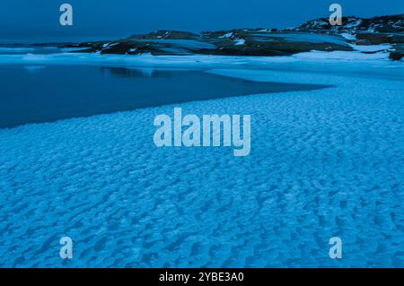 Un paysage côtier en hiver présente des eaux glacées et des terres enneigées, illuminées par une teinte bleue fraîche sous un ciel nocturne à Gothenburg, en Suède. Banque D'Images