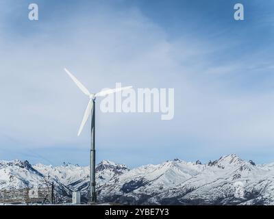 Une grande éolienne se dresse sur un fond de montagnes enneigées et un ciel bleu clair. La scène met en évidence le mélange de technologie et de n Banque D'Images