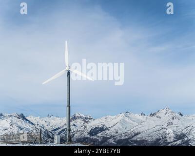 Une grande éolienne se dresse sur un fond de montagnes enneigées et un ciel bleu clair. La scène met en évidence le mélange de technologie et de n Banque D'Images