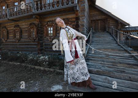 Une jeune fille avec une longue tresse blonde dans un costume folklorique, dans un village au milieu de l'architecture en bois, style slave Banque D'Images