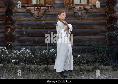 Une jeune fille avec une longue tresse blonde dans un costume folklorique, dans un village au milieu de l'architecture en bois, style slave Banque D'Images