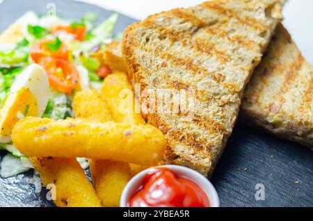 Une assiette de nourriture avec un sandwich, des bâtonnets de fromage et du ketchup. Le sandwich est coupé en deux et les frites sont dorées. Le ketchup est dans une petite tasse Banque D'Images