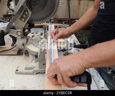 les mains mâles avec mètre ruban et crayon mesurent la longueur de la planche de bois sur l'établi avec la scie à onglet, marquage du matériel avant le traitement avec circulaire Banque D'Images