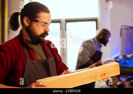 Menuisier portant des lunettes de sécurité tenant le bloc de bois dans les mains, mesurant la taille avant de commencer l'assemblage des meubles en studio. Menuisier en menuiserie se préparant à commencer à travailler sur un morceau de bois Banque D'Images