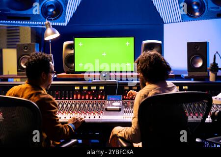 Équipe d'artistes mixant et mastering de la musique avec écran vert sur pc en studio professionnel, en utilisant la console audio avec des commutateurs et des curseurs dans la salle de contrôle. Chanteur et technicien du son créent des chansons. Banque D'Images