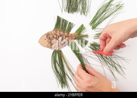 Personne coupant un bateau en forme de poisson avec des pommes de pin, des aiguilles de pin, des ciseaux rouges. Bricolage plat, mains de femme. Projet éducatif, activité enfants, créatif Banque D'Images