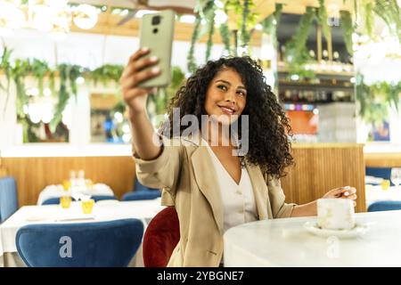 Femme latine heureuse et beauté avec de longs cheveux bouclés et des vêtements chics prenant selfie assis seul dans la cafétéria Banque D'Images