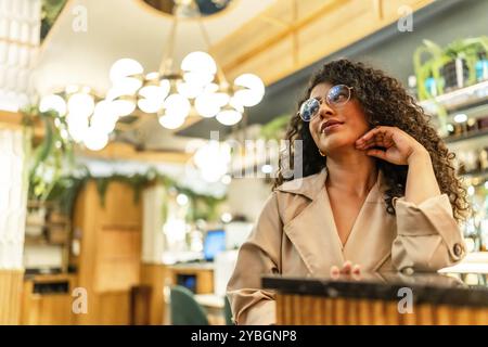 Élégante femme de beauté latine avec les cheveux bouclés attendant la main sur le menton dans une cafétéria moderne Banque D'Images