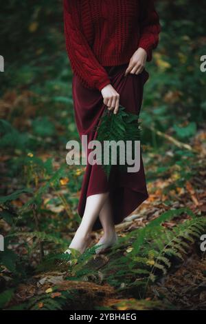 Une femme portant une robe Bordeaux se tient pieds nus dans un cadre de forêt sereine, entourée de fougères vertes vibrantes. Elle tient doucement une grande feuille, embodyi Banque D'Images