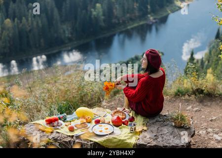 Une femme dans un pull rouge confortable est assis au bord d'un lac tranquille, entouré par un feuillage d'automne vibrant tout en profitant d'un pique-nique disposé sur une couverture avec divers Banque D'Images