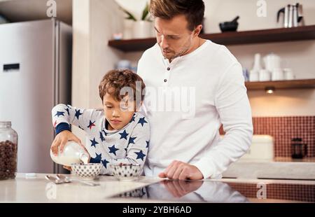 Lait, céréales et père avec enfant dans la cuisine pour le petit déjeuner à la maison le week-end pour la liaison. Heureux, porridge et papa aidant l'enfant garçon à préparer le matin Banque D'Images