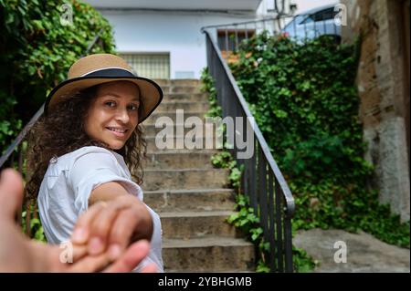 Une femme souriante dans un chapeau de paille tient une main, menant à de charmantes marches en plein air entourées d'une végétation luxuriante. La scène capture un sentiment d'aventure et Banque D'Images