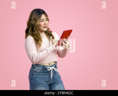 Jeune femme asiatique dans des vêtements décontractés, tenant et regardant une tablette. Portrait sur un fond rose simple avec lumière de studio. Vue latérale Banque D'Images