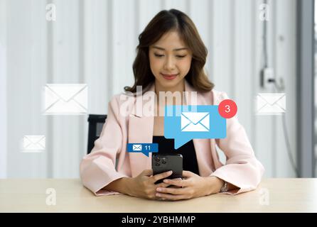 Femme d'affaires en costume rose assis au bureau dans le bureau, tapant sur le téléphone portable. Affichage des icônes d'e-mail et des notifications. Banque D'Images