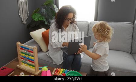 Mère travaillant sur un ordinateur portable pendant que son fils joue dans un salon moderne, soulignant un lien familial confortable à l'intérieur avec des jouets éducatifs autour d'eux, port Banque D'Images