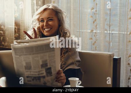 Touriste joyeux est assis dans un café, profitant d'une boisson chaude et lisant un journal, tout en gestant la paix avec ses doigts Banque D'Images