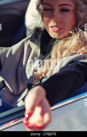 Une jeune femme aux longs cheveux blonds conduit une voiture décapotable par une journée ensoleillée. Sentir le vent dans ses cheveux et faire un geste joyeux par la fenêtre Banque D'Images