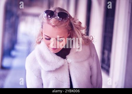 Femme blonde portant des lunettes de soleil et un manteau blanc marche dans un couloir extérieur avec des arcs, regardant vers le bas réfléchi Banque D'Images