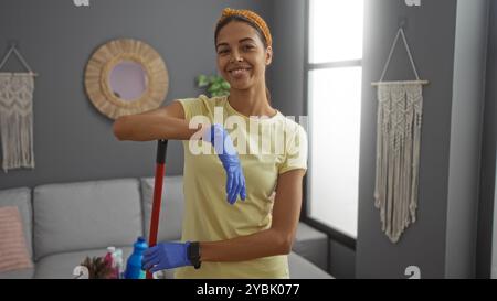 Jeune femme en gants nettoyant un salon moderne, souriant en toute confiance tout en tenant un balai près d'un canapé avec des produits de nettoyage Banque D'Images