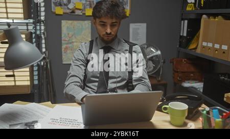 Un jeune homme arabe avec une barbe travaille sur un ordinateur portable dans le bureau d'un détective, entouré de dossiers et d'une carte. Banque D'Images