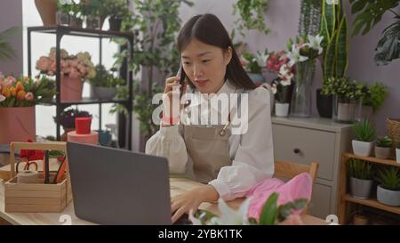Une femme chinoise fleuriste dans un tablier utilise un ordinateur portable et un téléphone à l'intérieur d'un magasin de fleurs entouré de bouquets et de plantes fraîches. Banque D'Images