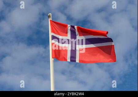 Un drapeau rouge frappant avec une croix bleue et blanche audacieuse flottant gracieusement dans la brise sous un ciel bleu clair orné de nuages blancs moelleux, s Banque D'Images