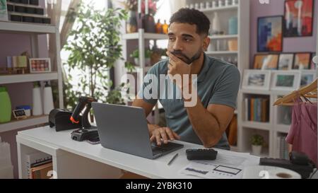 Jeune homme regardant réfléchi dans un magasin de décoration intérieure, assis à un bureau avec un ordinateur portable et divers outils d'emballage de produits et entouré d'un décor élégant Banque D'Images