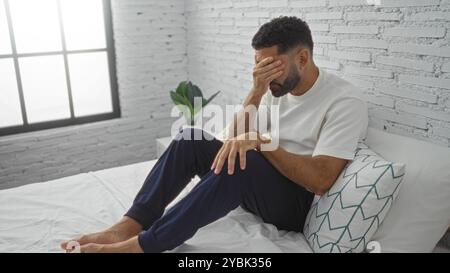 Le jeune homme est assis sur le lit dans une chambre moderne avec un mur de briques blanches recouvrant son visage, paraissant en détresse ou fatigué, avec une plante visible près de la fenêtre Banque D'Images