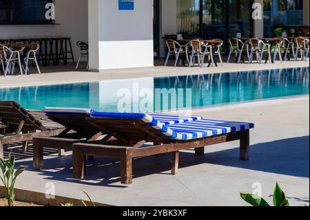 Sous un ciel bleu clair, des chaises longues aux rayures vibrantes se trouvent à côté d'une piscine étincelante, tandis que des tables et des chaises attendent les clients autour de la sérénité Banque D'Images