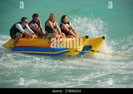 Les gens s'amusant sur un bateau de banane gonflable étant remorqué à grande vitesse derrière une côte. Banque D'Images