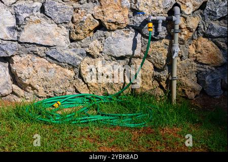 La lumière du soleil illumine un tuyau d'arrosage vert soigneusement enroulé sur l'herbe à côté d'un mur de pierre robuste, créant un environnement extérieur paisible. Banque D'Images