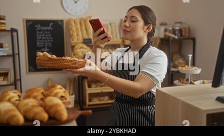 Femme prenant une photo dans une boulangerie remplie de pain tout en tenant un panier, entourée de pâtisseries et de produits de boulangerie, montrant un commerce intérieur Banque D'Images