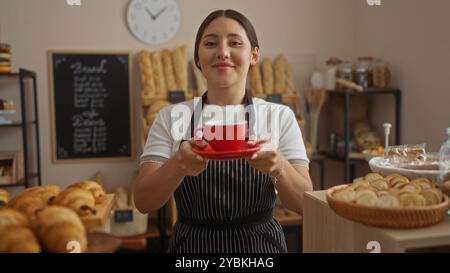 Jeune femme hispanique souriante tenant une tasse rouge dans une boulangerie confortable avec du pain et des pâtisseries en arrière-plan Banque D'Images