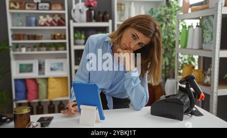 Femme travaillant dans le magasin de décoration à la maison en utilisant une tablette avec une expression réfléchie entourée de diverses décorations Banque D'Images