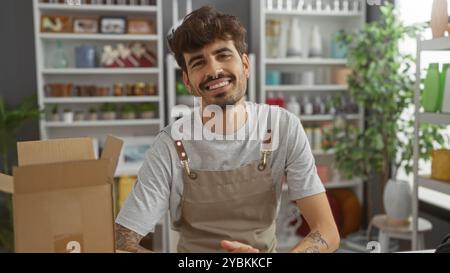 Jeune homme hispanique à la barbe souriant dans un magasin de décoration entouré de diverses décorations et plantes Banque D'Images