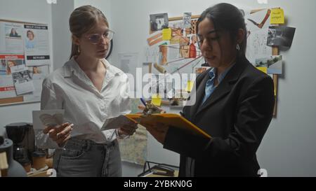 Deux femmes enquêtrices analysent les preuves dans un bureau de poste de police encombré, façonné par les exigences de leur profession. Banque D'Images