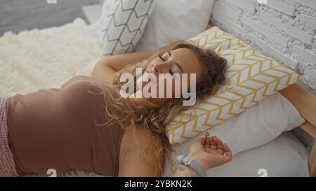 Femme se relaxant sur le lit à la maison avec des oreillers confortables, montrant un jeune adulte attrayant blond reposant paisiblement à l'intérieur dans une chambre confortable Banque D'Images