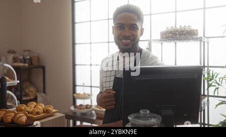 Jeune homme travaillant dans la boulangerie souriant à la caisse avec des pâtisseries affichées dans un cadre intérieur confortable Banque D'Images