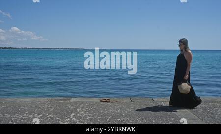 Une jeune femme hispanique marche le long des eaux bleues de porto cesareo, salento par une journée ensoleillée, portant un sac de paille et portant une robe noire. Banque D'Images