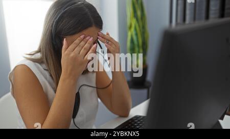 Une jeune femme portant un casque semble stressée lorsqu'elle est assise à un bureau avec un ordinateur devant elle. Banque D'Images